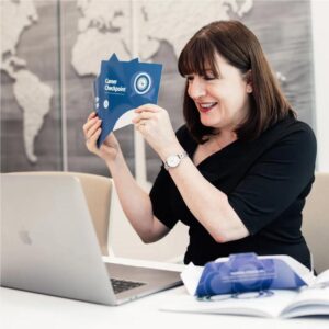Antoinette Oglethorpe sits at a desk smiling whilst holding up a card from her Career Conversation Toolkit titled 