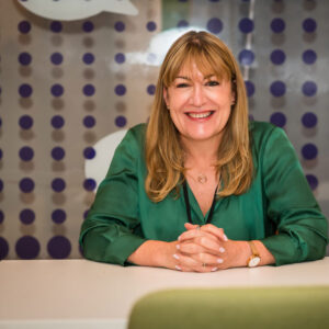 Antoinette Oglethorpe shares her exceptional client experiencxe stories and case studies, image of Antoinette with long honey blonde hair, wearing a green blouse, sits at a white table smiling. The background features a glass wall with purple dots and white speech bubble shapes.