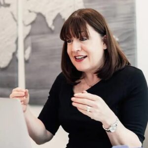 A woman with shoulder-length brown hair sits at a desk, smiling and gesturing with her hands whilst looking at a laptop. A world map artwork is visible on the wall behind her.